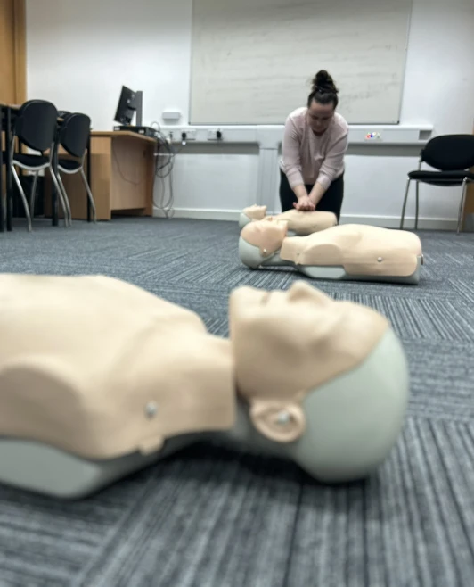 A CPR mannequin lies in focus on the floor, with a woman in the background performing chest compressions as part of a first aid training session. A CPR mannequin lies in focus on the floor, with a woman in the background performing chest compressions as part of a first aid training session.
